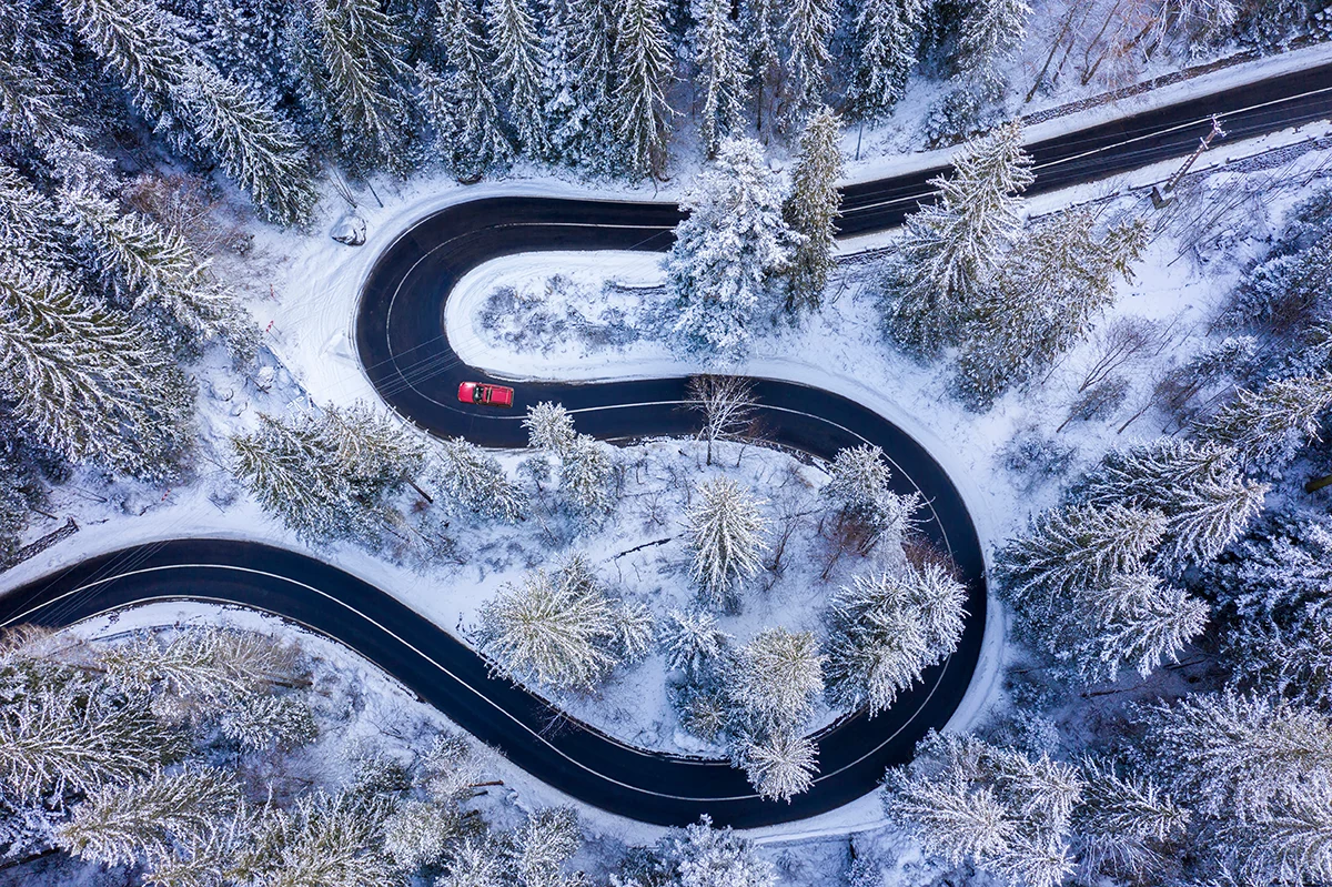 A red car driving a winding canyon road flanked by snow and pine trees.