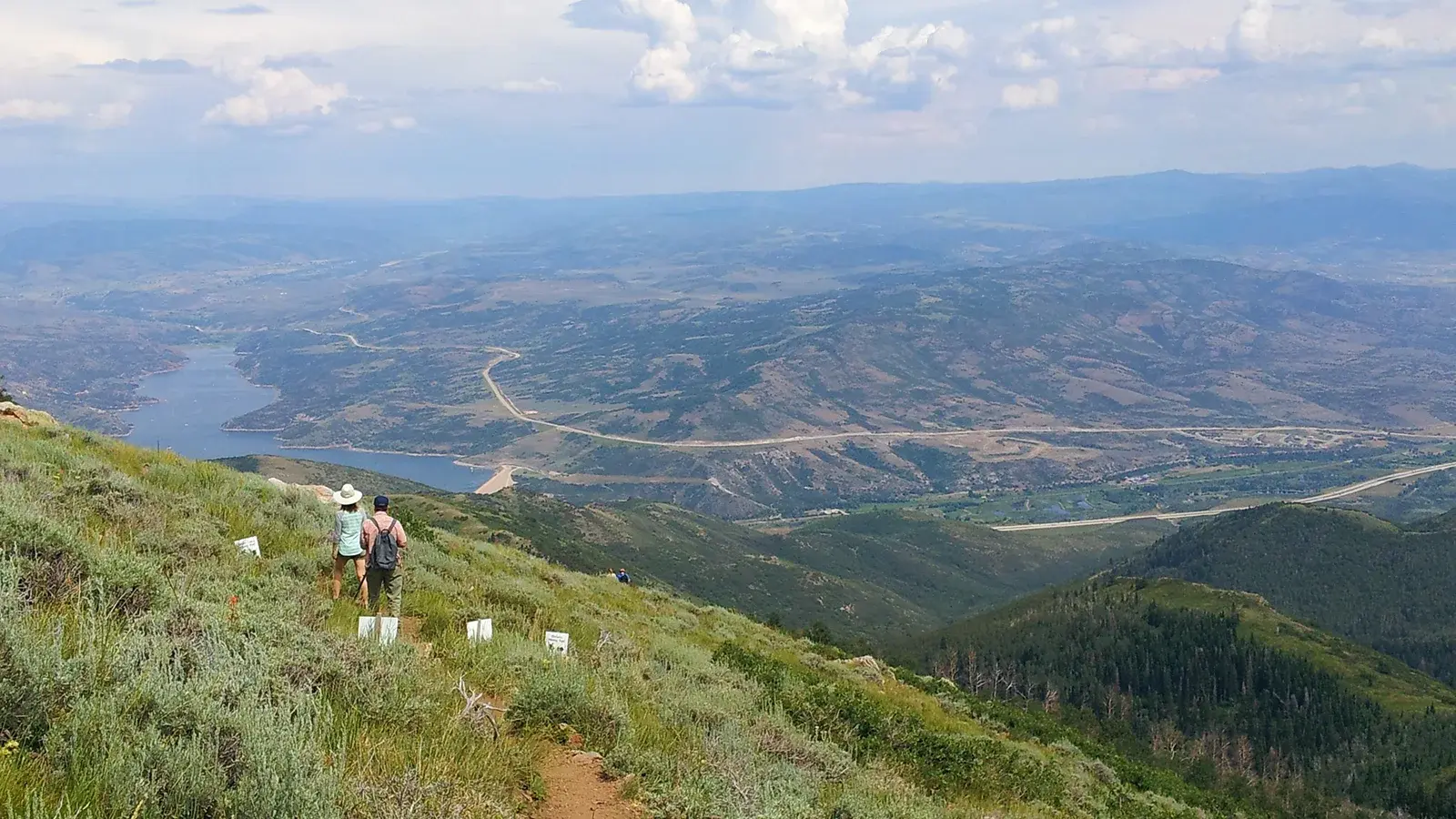 A grassy mountain with two hikers in the foreground with Deer Valley in the background