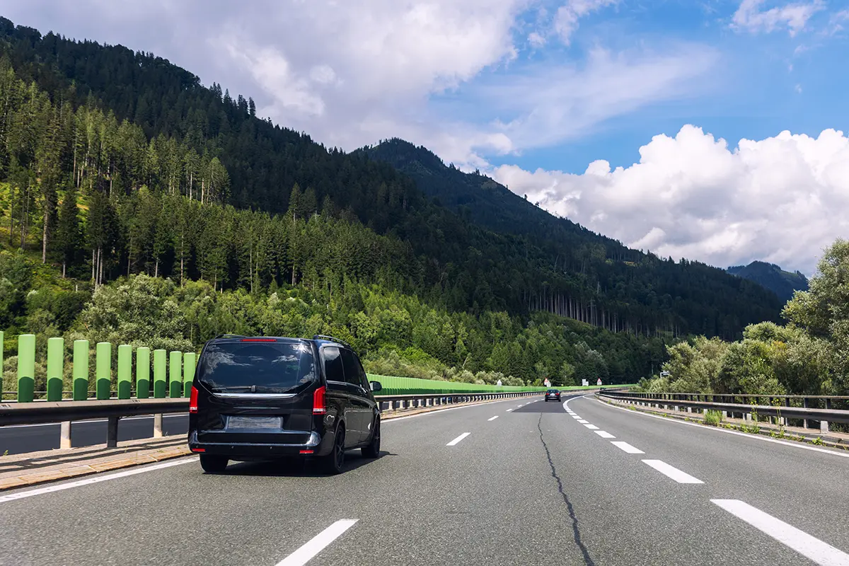 Dark car driving a two-lane mountain road flanked by a guardrail, pine trees, and hills