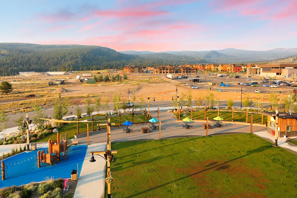 An aerial view of outdoor space that consists of grass playing area, playground with climbing structures, a patio, and a shopping area in the background