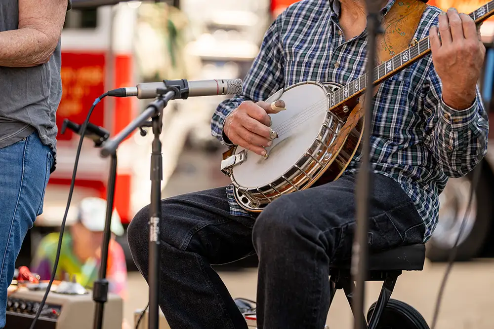 Close-up of banjo player's hands plucking strings at outdoor performance with microphone nearby amplifying music. Captures bluegrass, folk music traditions, string instruments, and live music festival