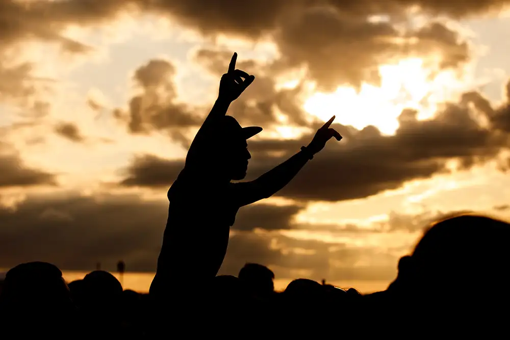 A man in a cap and raised arms takes pleasure at an outdoor music festival. Black silhouette on sunset.